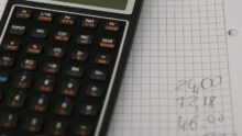 Un main, un cahier et une calculatrice. La main reporte les résultats des calculs sur le cahier