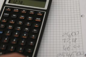 Un main, un cahier et une calculatrice. La main reporte les résultats des calculs sur le cahier