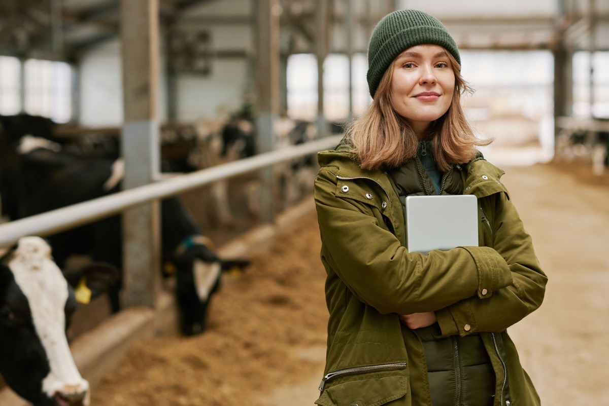 Portrait d'une jeune femme agronome avec une tablette debout dans une stabulation avec des vaches - Illustration Le contrôle fin d’engagements JA assoupli