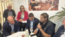 Un groupe de personne autour d'une table en train d'écouter un homme qui parle avec un micro à la main