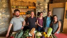 Un groupe de personne dans une pièce avec des murs de pierre, avec des cageots remplis de légumes posés sur une table devant eux