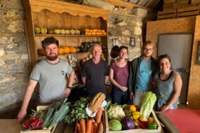 Un groupe de personne dans une pièce avec des murs de pierre, avec des cageots remplis de légumes posés sur une table devant eux