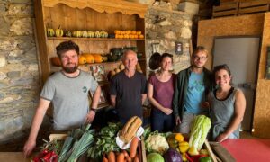 Un groupe de personne dans une pièce avec des murs de pierre, avec des cageots remplis de légumes posés sur une table devant eux