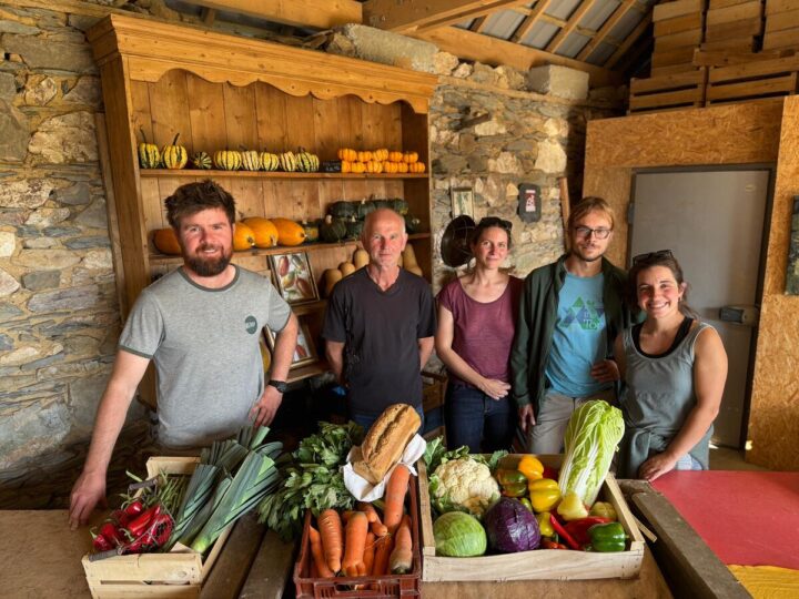 Un groupe de personne dans une pièce avec des murs de pierre, avec des cageots remplis de légumes posés sur une table devant eux