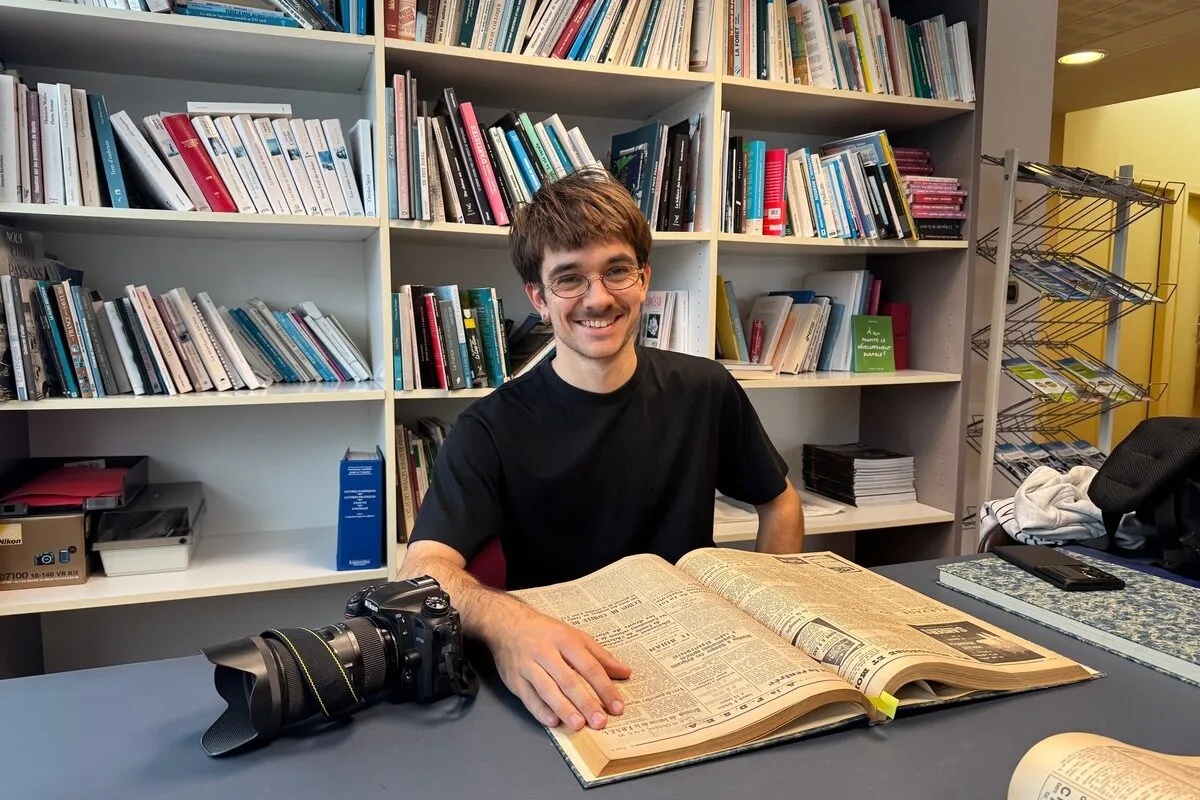 Un jeune photographe consulte un vieux journal d'archives devant des étagères remplies de livres. - Illustration Sur les traces du remembrement breton