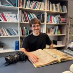 Un jeune photographe consulte un vieux journal d'archives devant des étagères remplies de livres.
