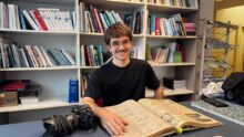 Un jeune photographe consulte un vieux journal d'archives devant des étagères remplies de livres.