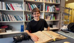 Un jeune photographe consulte un vieux journal d'archives devant des étagères remplies de livres.
