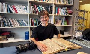 Un jeune photographe consulte un vieux journal d'archives devant des étagères remplies de livres.