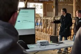 Des personnes dans un bâtiment d'élevage bovin avec en premier plan le dos d'un homme qui regarde un écran d'ordinateur