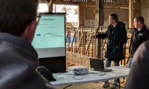 Des personnes dans un bâtiment d'élevage bovin avec en premier plan le dos d'un homme qui regarde un écran d'ordinateur