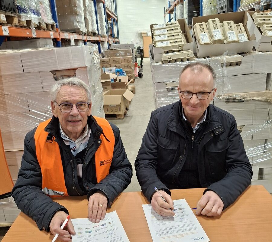 Deux hommes souriants assis à une table signent un contrat avec des palettes d'oeufs derrière - Illustration 180 000 œufs offerts aux Banques alimentaires de l’Ouest