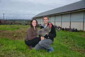 Une femme et un homme accroupis devant un bâtiment d'élevage de volailles avec une poule Coucou de Rennes dan les bras