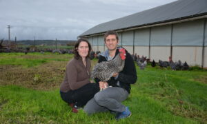 Une femme et un homme accroupis devant un bâtiment d'élevage de volailles avec une poule Coucou de Rennes dan les bras