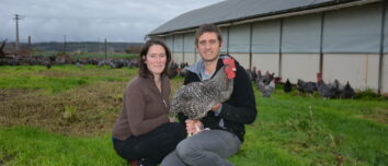 Une femme et un homme accroupis devant un bâtiment d'élevage de volailles avec une poule Coucou de Rennes dan les bras