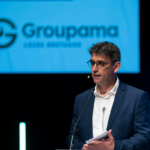 Un homme avec un micro casque à une tribune un écran projette le logo groupama derrière