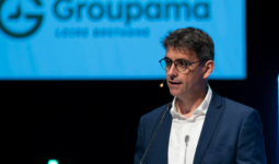 Un homme avec un micro casque à une tribune un écran projette le logo groupama derrière