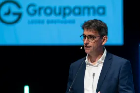 Un homme avec un micro casque à une tribune un écran projette le logo groupama derrière