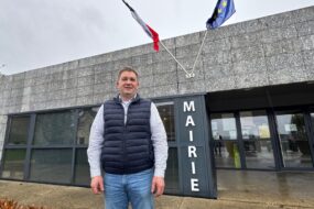 Un homme devant une mairie présentant les drapeaux de la France et de l'Europe.
