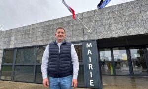 Un homme devant une mairie présentant les drapeaux de la France et de l'Europe.