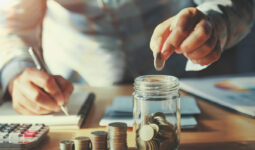 Un homme qui écrit sur un cahier de la main doite et qui met des pièces dans un bocal en verre de la main gauche