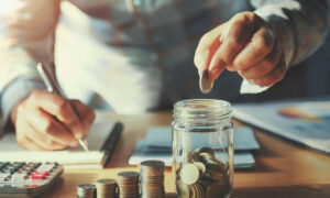 Un homme qui écrit sur un cahier de la main doite et qui met des pièces dans un bocal en verre de la main gauche