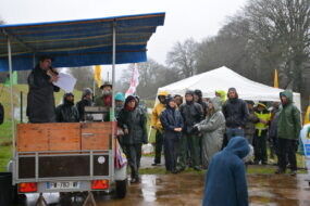 Des manifestants écoutent, sous la pluie, une personne avec un micro installée sur une remorque couverte
