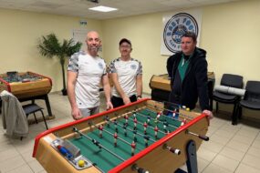 Trois hommes autour d'un baby-foot dans une salle avec plusieurs autres baby-foot