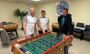 Trois hommes autour d'un baby-foot dans une salle avec plusieurs autres baby-foot