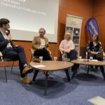 Des hommes et femmes assis derrière des tables basses dans une salle de réunion. Un homme parle dans un micro