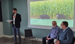 2 hommes assis sur une chaise et un 3ème debout avec un micro devant un grand écran qui projette des sangliers dans un champ de maïs