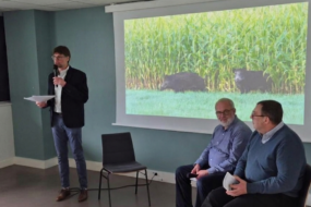 2 hommes assis sur une chaise et un 3ème debout avec un micro devant un grand écran qui projette des sangliers dans un champ de maïs