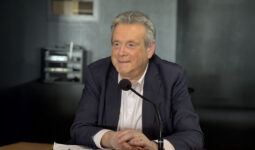 Un homme souriant d'une soixantaine d'années assis à une tribune derrière un micro