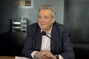 Un homme souriant d'une soixantaine d'années assis à une tribune derrière un micro