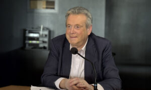 Un homme souriant d'une soixantaine d'années assis à une tribune derrière un micro