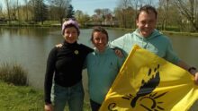 un homme et deux femmes au bord d'une rivière avec un drapeau de la Confédération Paysanne