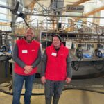 Deux éleveurs habllés d'une veste rouge devant leur manège de traite 28 postes de marque DeLaval