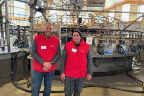 Deux éleveurs habllés d'une veste rouge devant leur manège de traite 28 postes de marque DeLaval