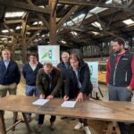 Six hommes dont deux en train de signer des documents sur une table posée dans un bâtiment agricole