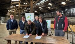 Six hommes dont deux en train de signer des documents sur une table posée dans un bâtiment agricole