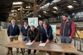 Six hommes dont deux en train de signer des documents sur une table posée dans un bâtiment agricole