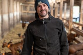 Un homme souriant avec un bonnet au mileu d'un troupeau de chèvres dans un bâtiment d'élevage