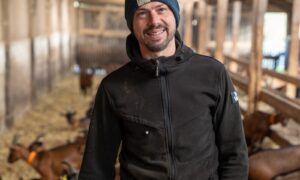 Un homme souriant avec un bonnet au mileu d'un troupeau de chèvres dans un bâtiment d'élevage