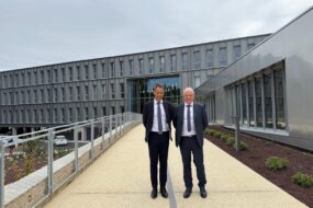 Deux hommes en costume sombre et cravate devant un bâtiment gris