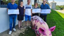Des personnes avec des grands chèques dans les mains devant un bâtiment d'élevage avec un cochon devant eux