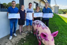 Des personnes avec des grands chèques dans les mains devant un bâtiment d'élevage avec un cochon devant eux