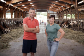 Couple de jeunes agriculteurs posant dans sa stabulation avec trois jeunes enfants jouant à l'arrière-plan