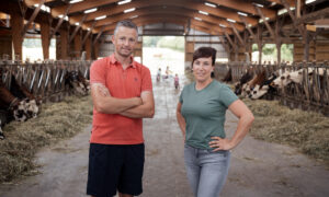 Couple de jeunes agriculteurs posant dans sa stabulation avec trois jeunes enfants jouant à l'arrière-plan