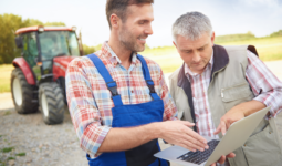 Deux hommes regardent un écran d'ordinateur portable au milieu d'une cour de ferme avec un tracteur en arrière plan