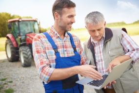 Deux hommes regardent un écran d'ordinateur portable au milieu d'une cour de ferme avec un tracteur en arrière plan